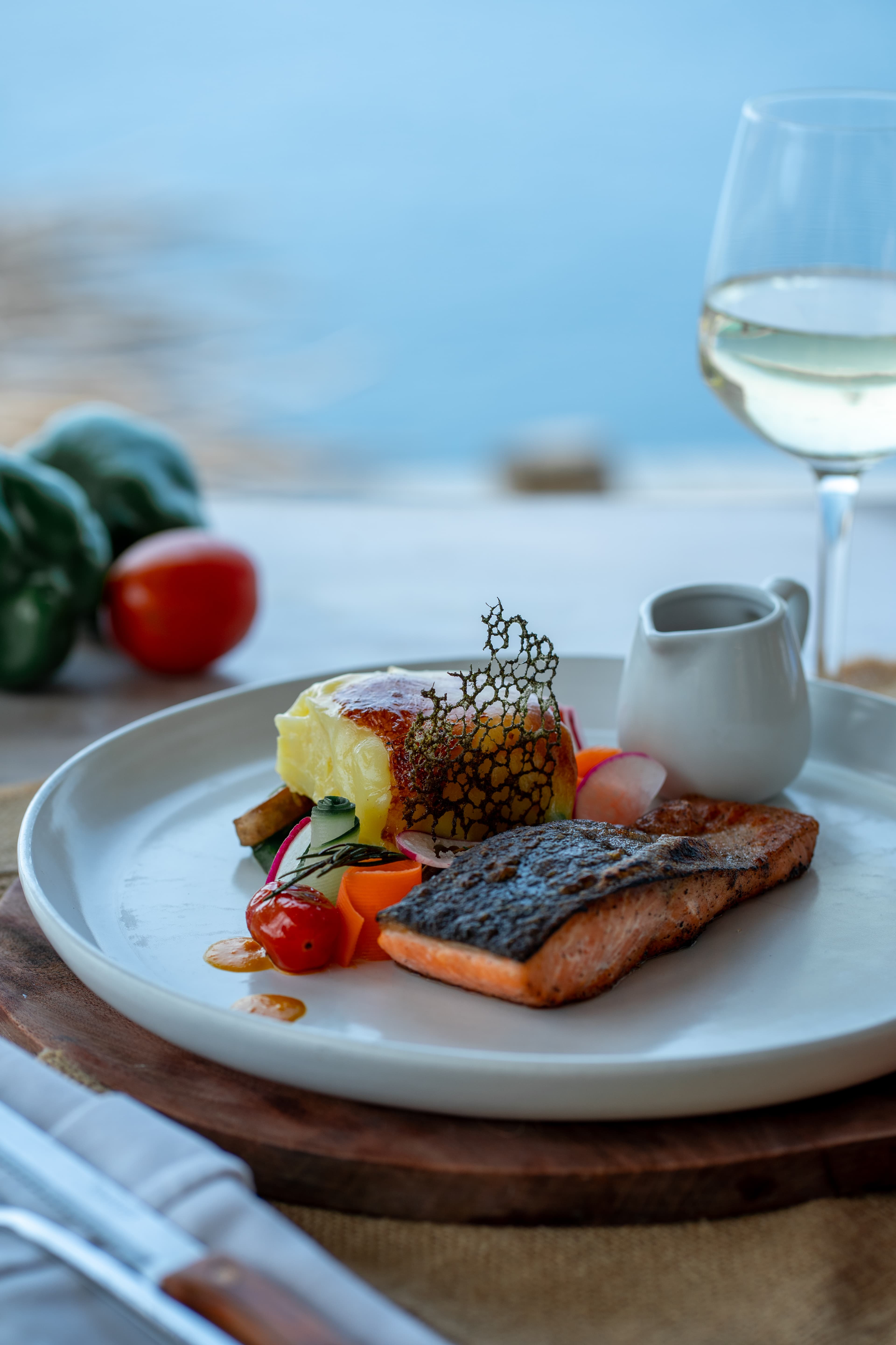Pan-seared salmon with potato gratin and fresh vegetables overlooking a calm blue ocean.