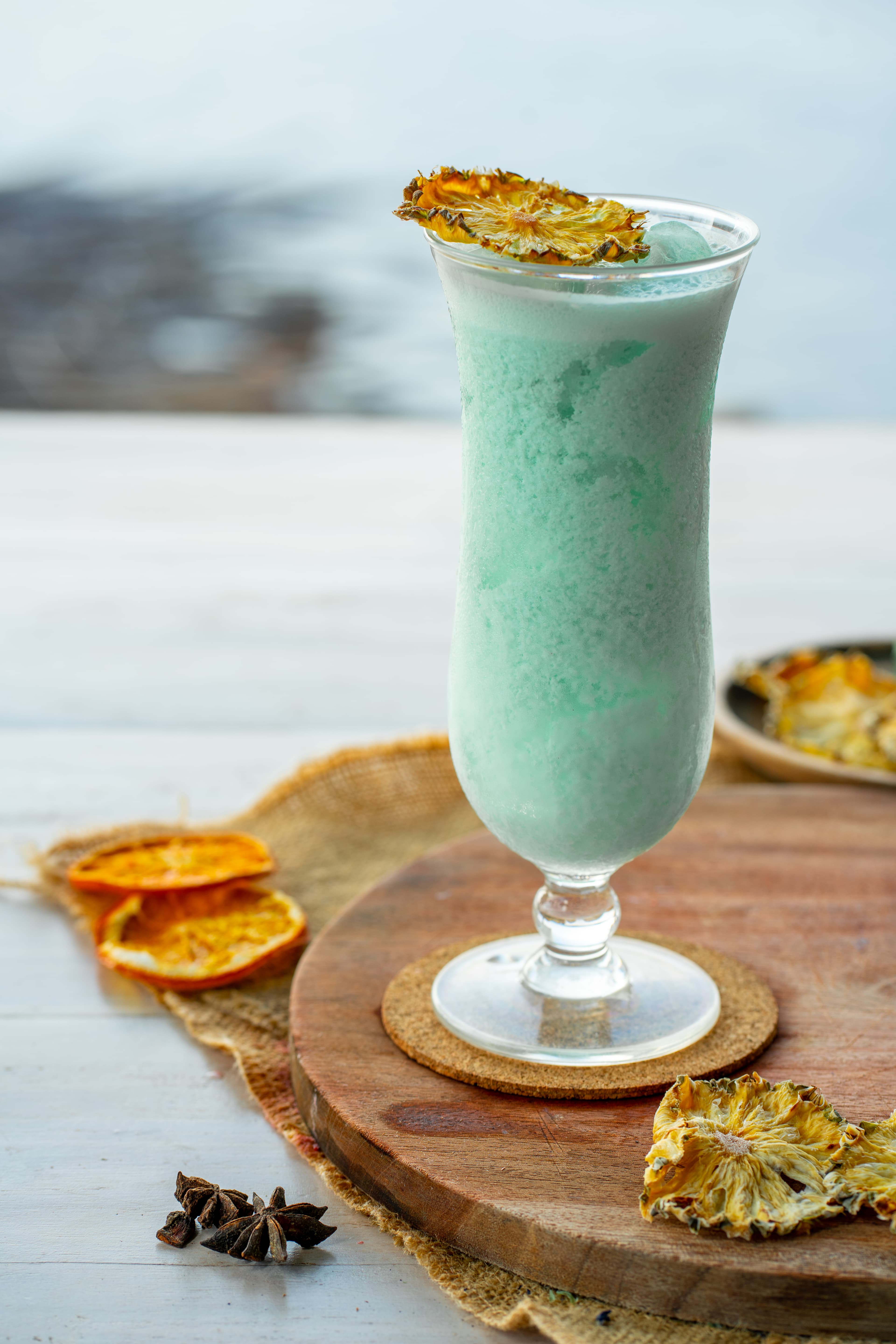 Light green frozen cocktail in tall glass garnished with dried pineapple on a wooden board.