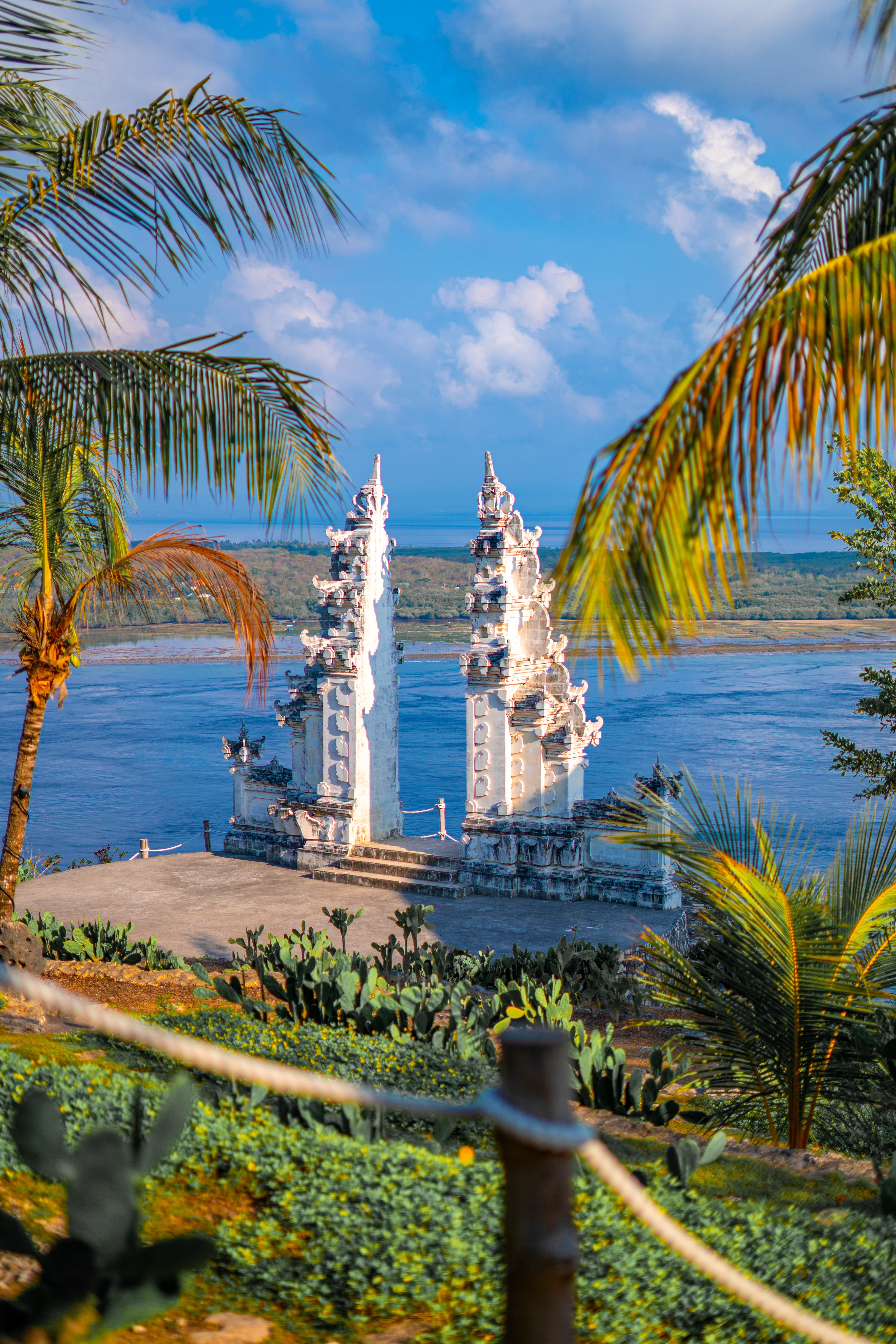 White Balinese split gate overlooking the ocean, framed by tropical palm trees and green cacti.