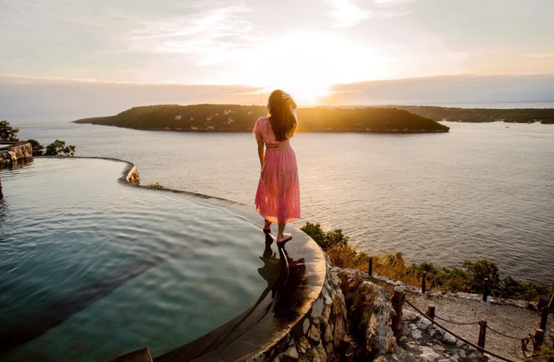 Woman in pink dress stands on infinity pool edge overlooking a sunset and distant island.