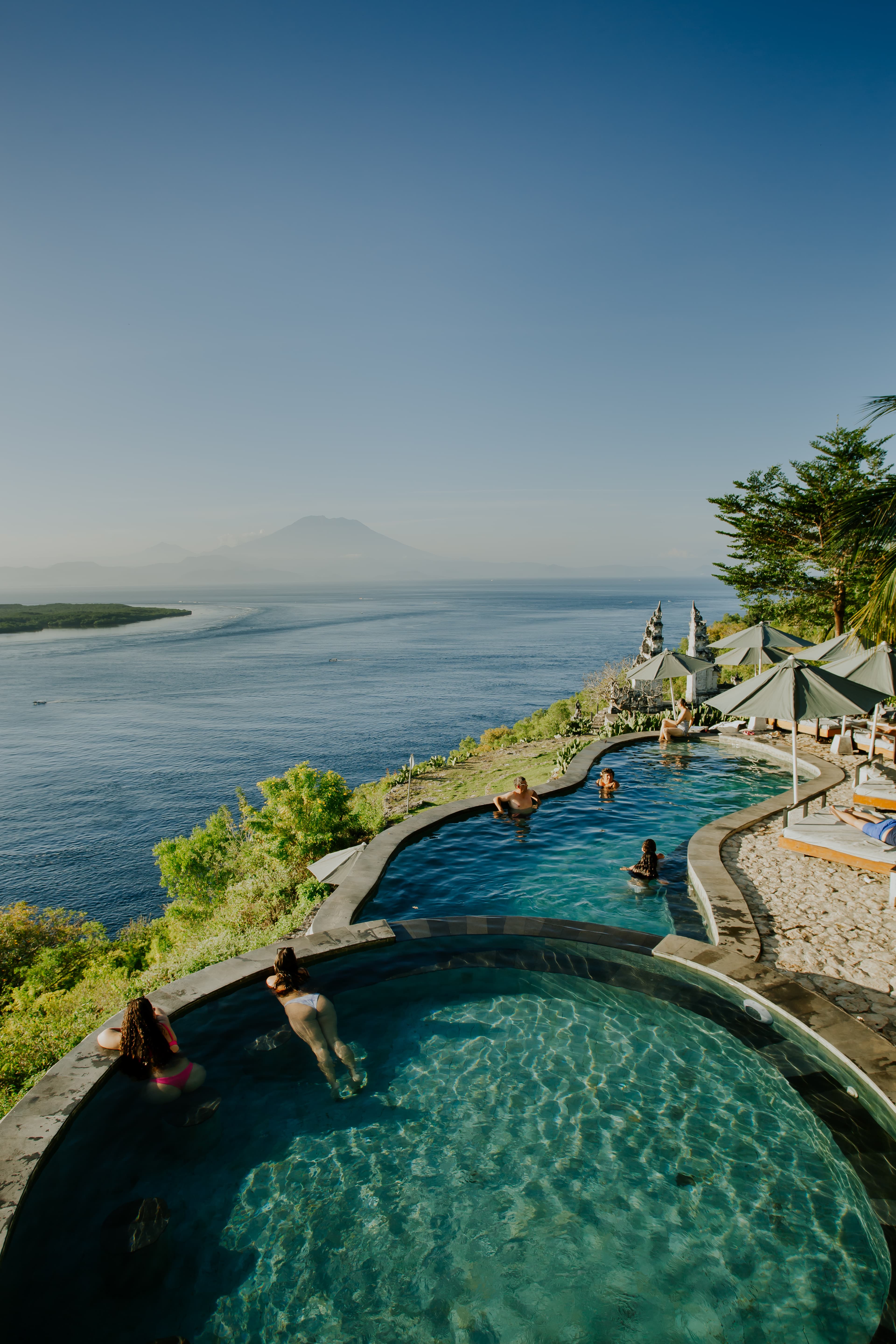 People swimming in tiered infinity pools overlooking the ocean and a distant mountain.