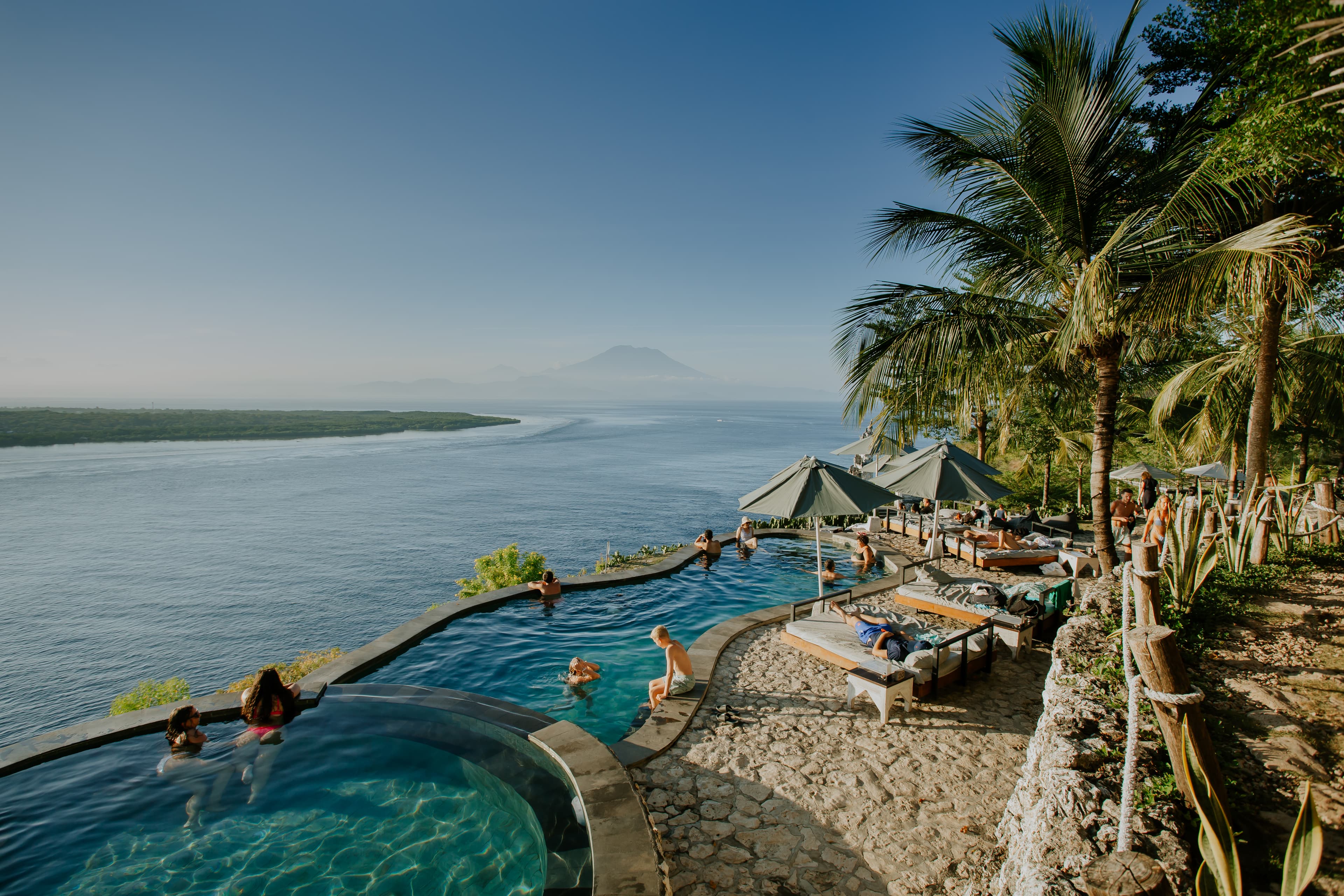 People relax in a tropical infinity pool overlooking the ocean and a distant mountain.
