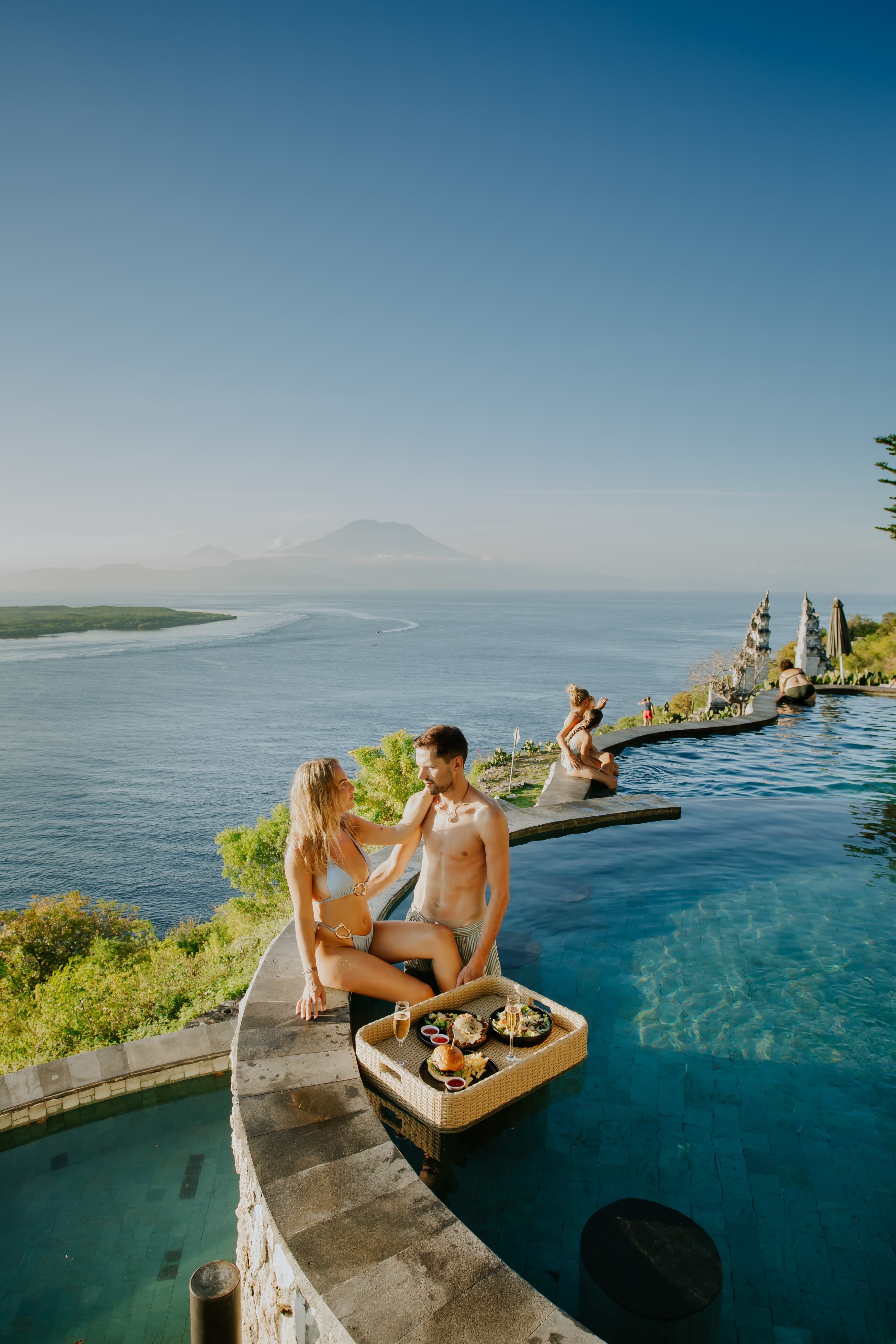 Couple enjoys a floating breakfast in an infinity pool overlooking the ocean and distant mountains.