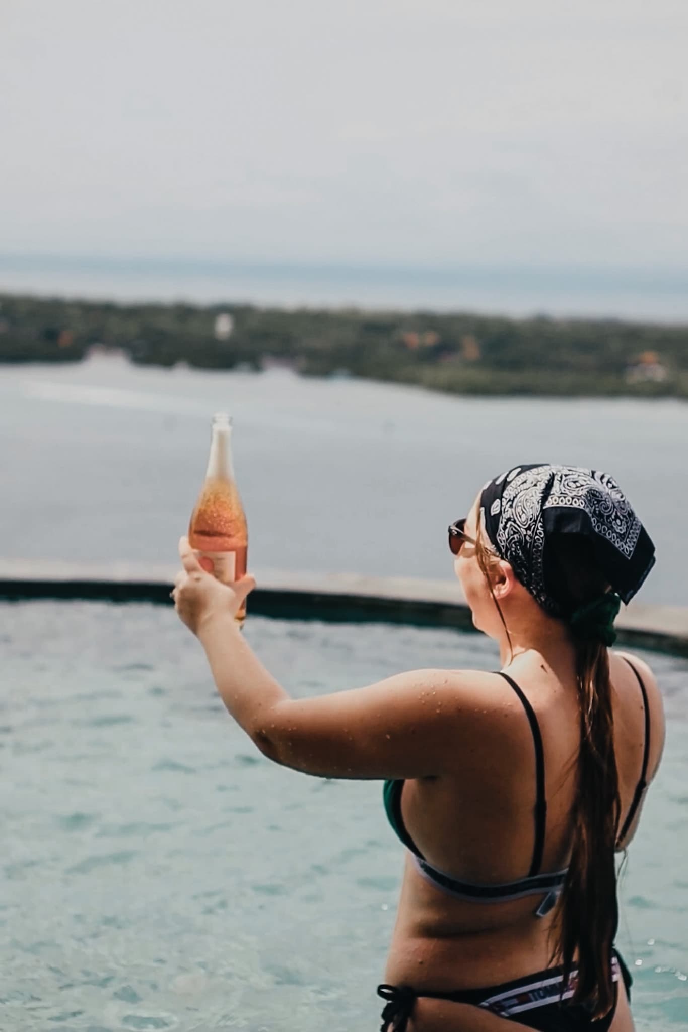 Woman in a bandana holds a bottle of wine in an infinity pool overlooking a coastal landscape.