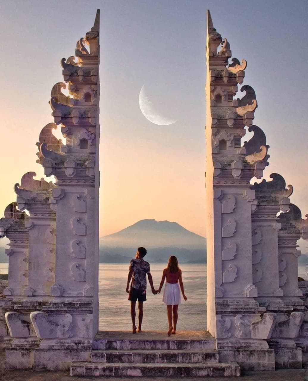 Couple holding hands between ornate Balinese gates overlooking a mountain and large crescent moon.