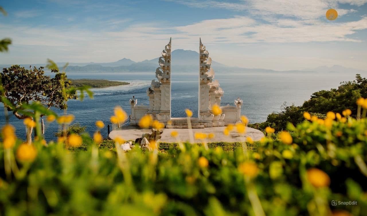 White Balinese split gate overlooking the ocean with a distant mountain and yellow flowers.