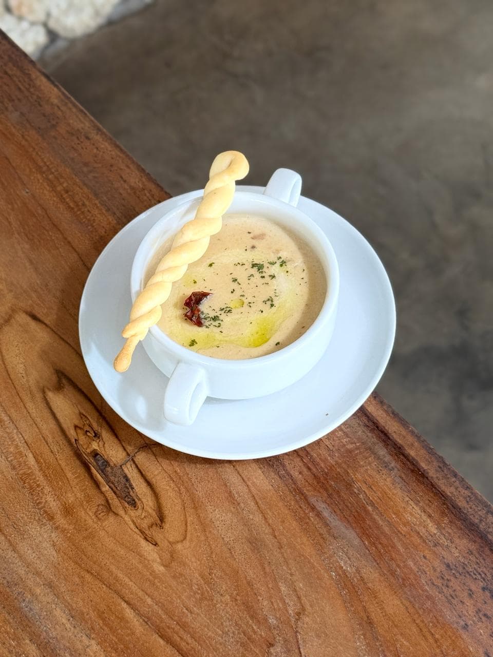 Creamy soup in a white bowl with a twisted breadstick, garnished with oil and herbs.