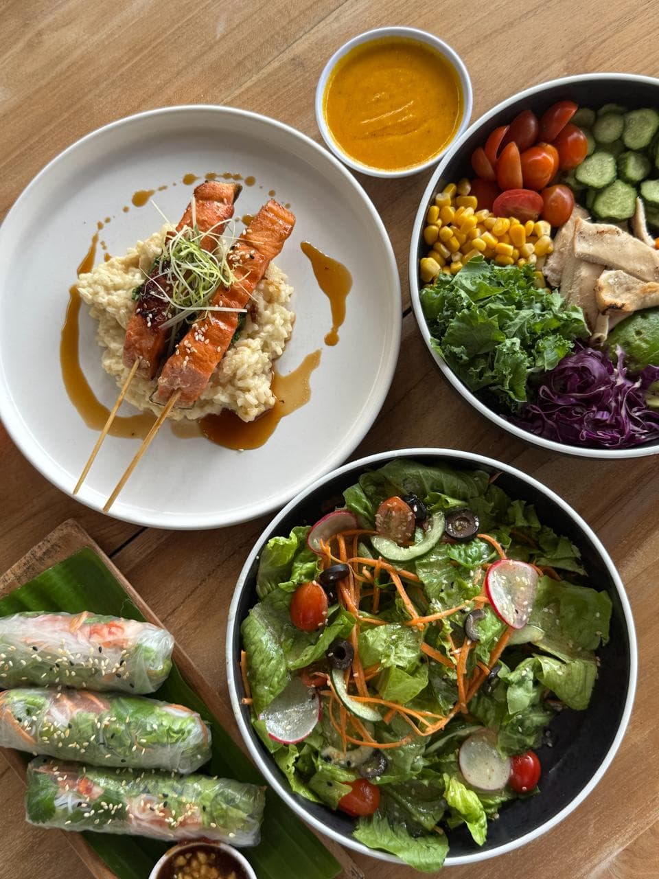 Grilled salmon skewers on risotto, a colorful vegetable bowl, garden salad, and spring rolls.