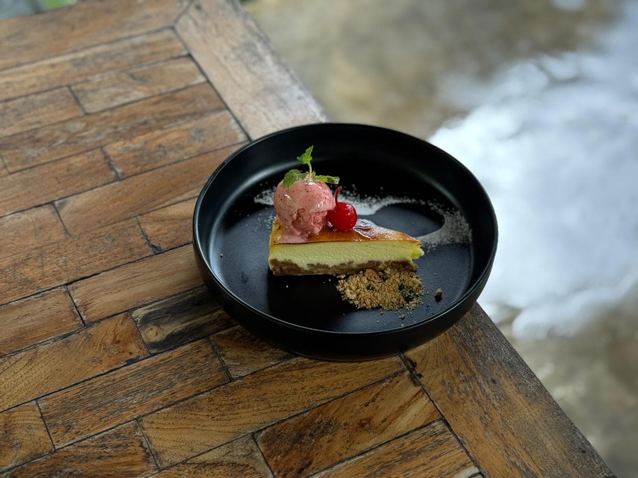 Cheesecake slice topped with pink sorbet, a cherry, and mint on a black plate.